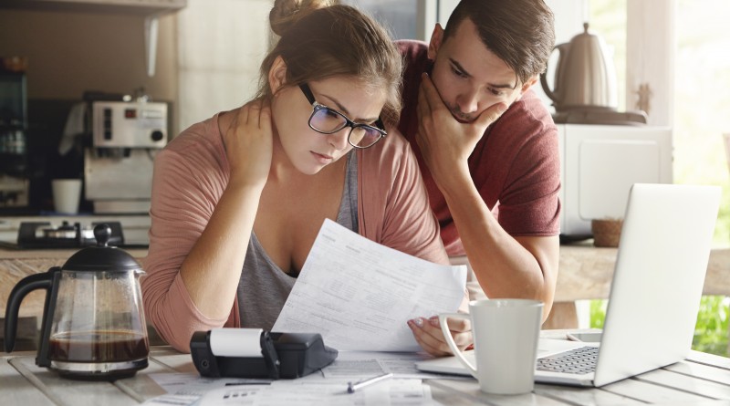Young Caucasian family having debt problems, not able to pay out their loan. Female in glasses and brunette man studying paper form bank while managing domestic budget together in kitchen interior